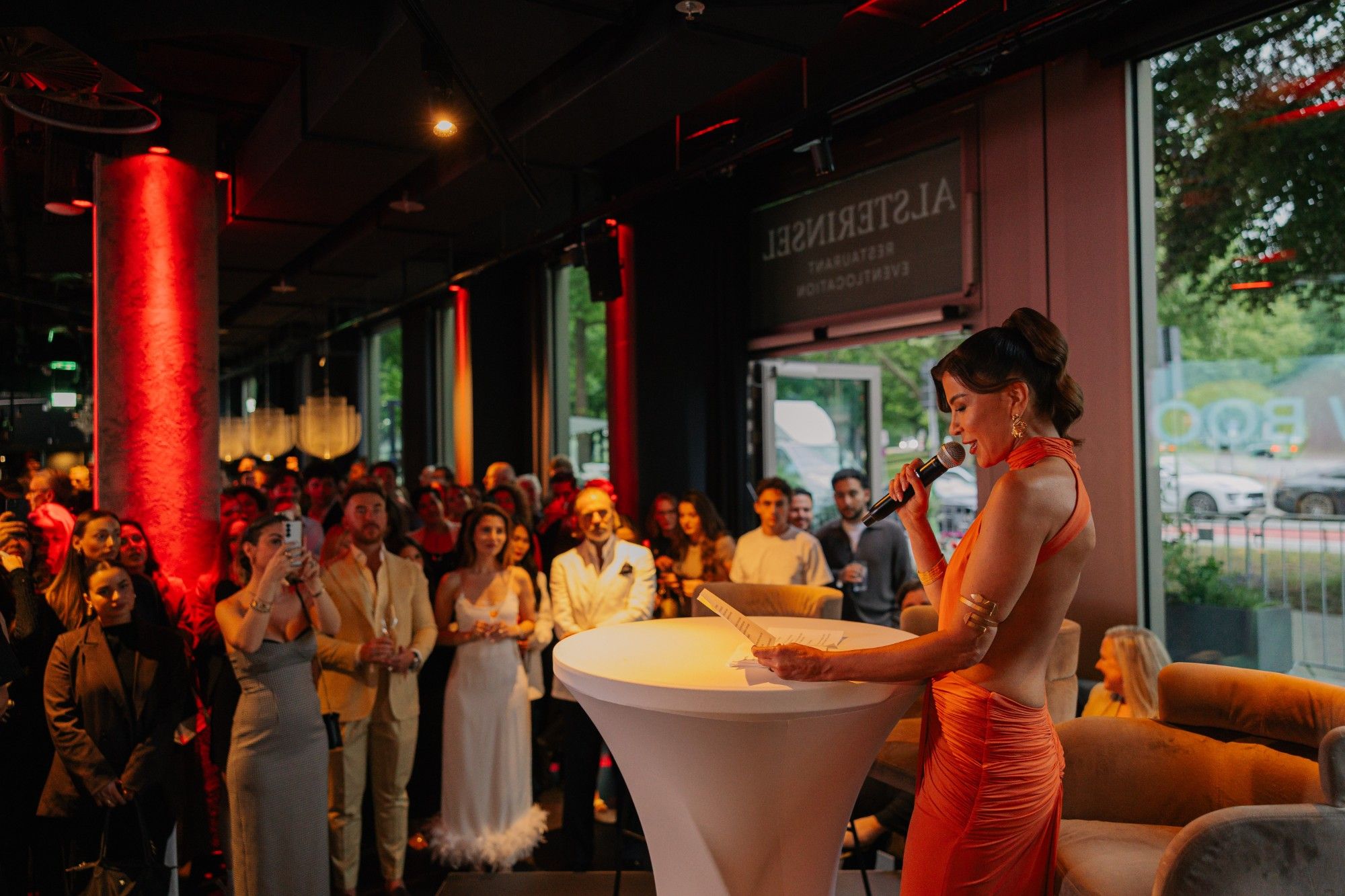 Gastgeberin in orangefarbenem Kleid hält eine Rede, die Gäste hören zu.