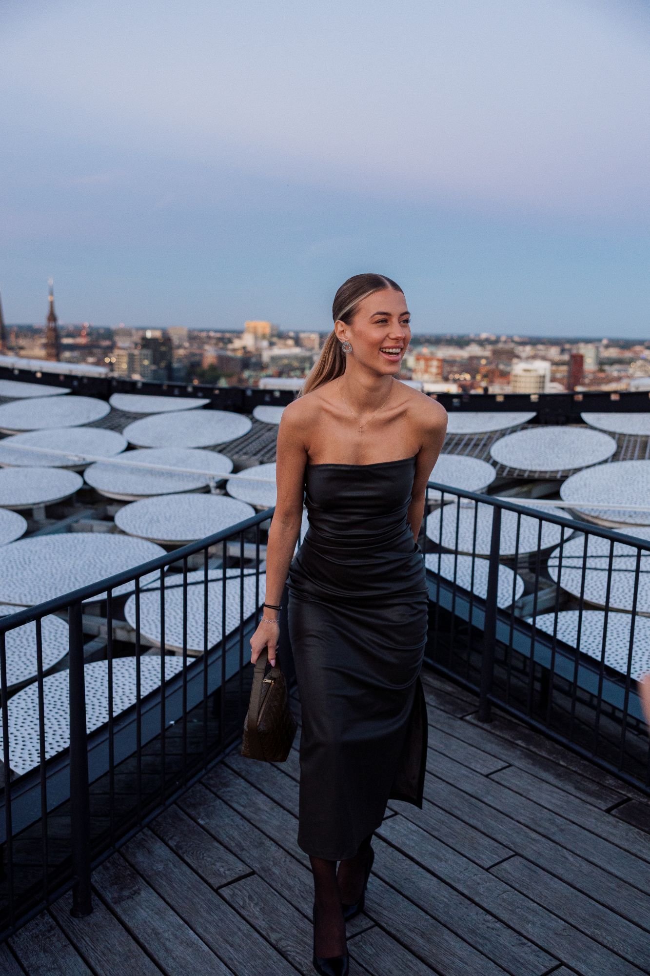 Gast in schwarzem Kleid lachend auf der Dachterrasse, dahinter die Stadt.