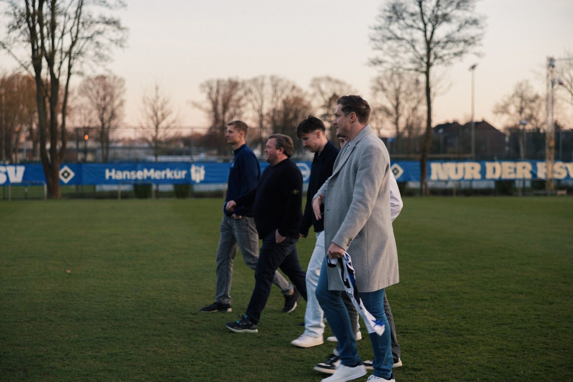 Eine Gruppe Männer geht im Gespräch über den Rasen, dahinter HSV-Banner.