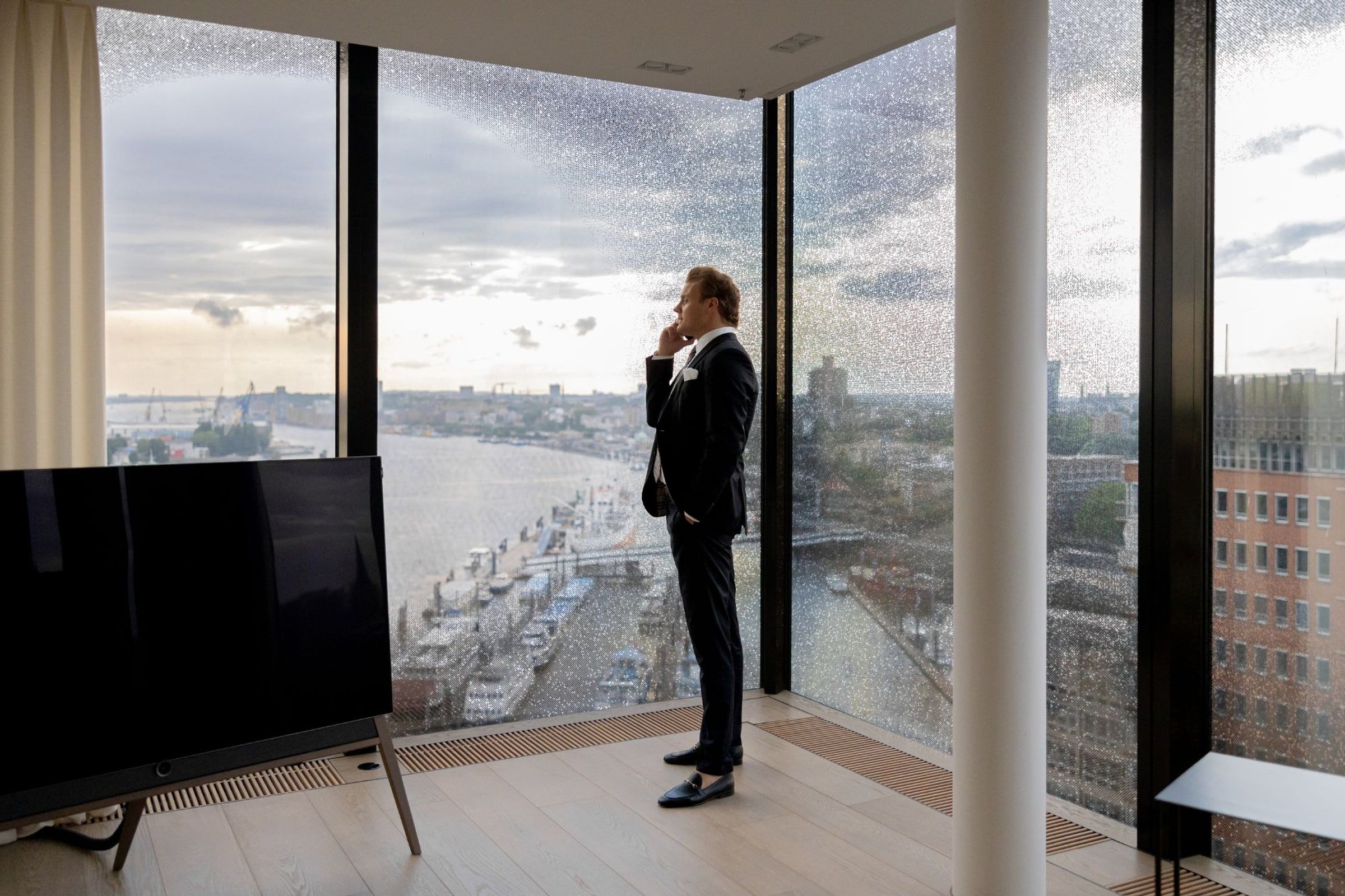 Geschäftsmann im Anzug telefoniert an einer bodentiefen Fensterfront hoch über dem Hamburger Hafen.