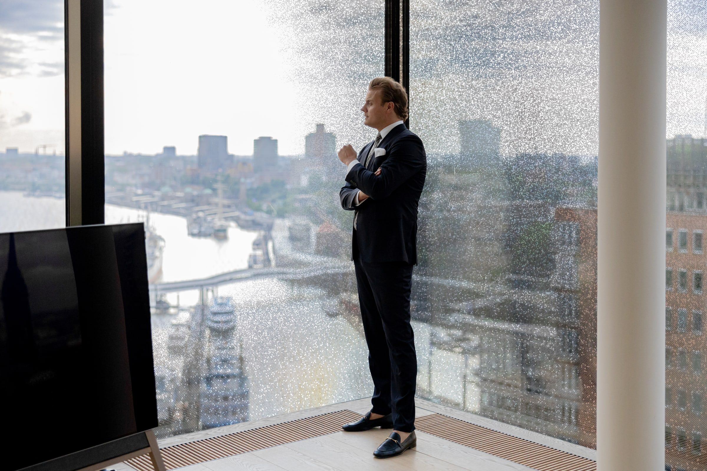 Björn Meins im Anzug an einer bodentiefen Fensterfront mit Blick über den Hafen.