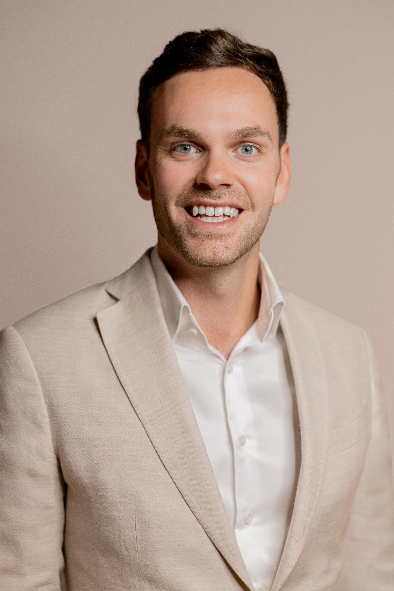 Portrait eines Mitarbeiters in beigem Sakko vor neutralem Hintergrund.